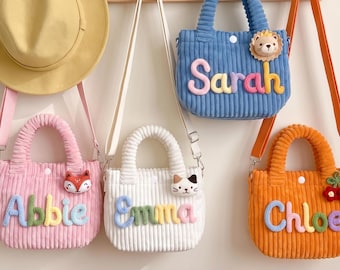 May include: A yellow hat and four corduroy handbags in pink, white, blue, and orange. Each bag features a plush animal or fruit accent and a colorful name: Abbie, Emma, Sarah, and Chloe. The bags have top handles and shoulder straps.
