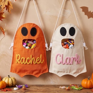 May include: Two ghost-shaped fabric treat bags hanging from metal hooks. The orange bag features the name Rachel in yellow yarn, and the cream bag features the name Clark in pink yarn. Both have black felt eyes and a central opening filled with candy.