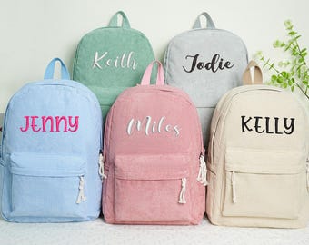 May include: Five corduroy backpacks in pastel colors: light blue, green, pink, gray, and cream. Each backpack has a front pocket and a name embroidered on the front: Jenny, Keith, Miles, Jodie, and Kelly. The backpacks have a top handle and adjustable straps.