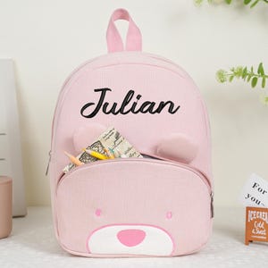 May include: Pink corduroy backpack with a bear face design and the name "Julian" in black script. The backpack has a front pocket, bear ears, and a top handle. Pencils and paper are visible in the pocket.