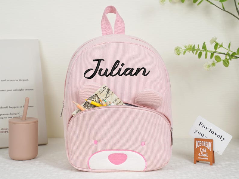May include: Pink corduroy backpack with a bear face design and the name "Julian" in black script. The backpack has a front pocket, bear ears, and a top handle. Pencils and paper are visible in the pocket.