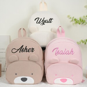 May include: Three corduroy backpacks in beige, white, and pink, each with a bear face design. The backpacks have the names "Asher", "Wyatt", and "Isaiah" embroidered on them. The bags have a top handle and adjustable straps.