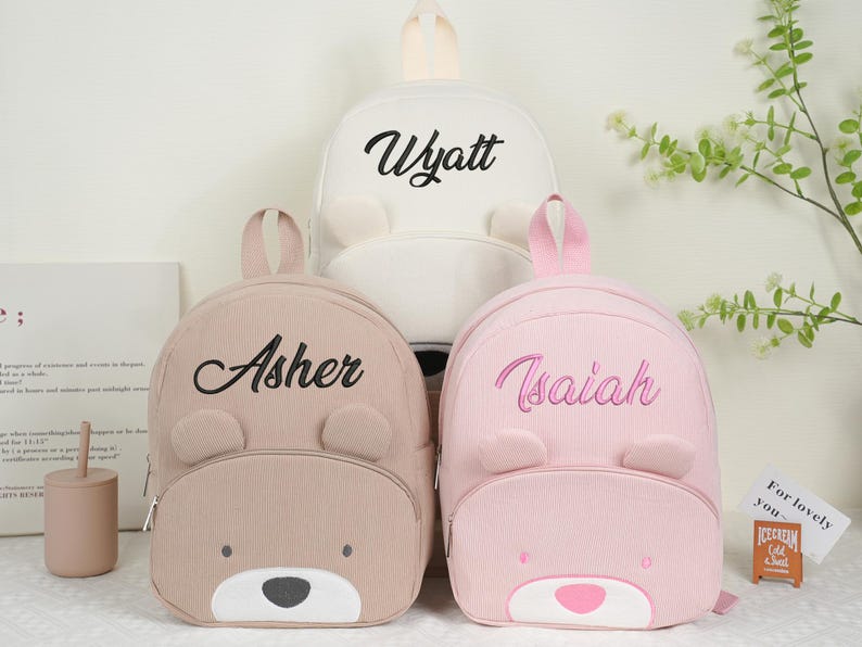 May include: Three corduroy backpacks in beige, white, and pink, each with a bear face design. The backpacks have the names "Asher", "Wyatt", and "Isaiah" embroidered on them. The bags have a top handle and adjustable straps.