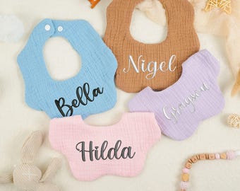 May include: Four personalized baby bibs with different names embroidered on them. The bibs are in blue, brown, pink, and purple. The names are Bella, Nigel, Grayson, and Hilda.