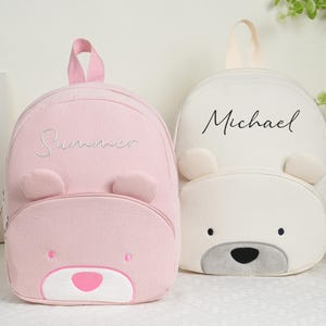 May include: Two children's backpacks, one pink and one white, each with a bear face design. The pink backpack has the name "Summer" embroidered on it, while the white one has "Michael". Both have bear ears and a top handle.