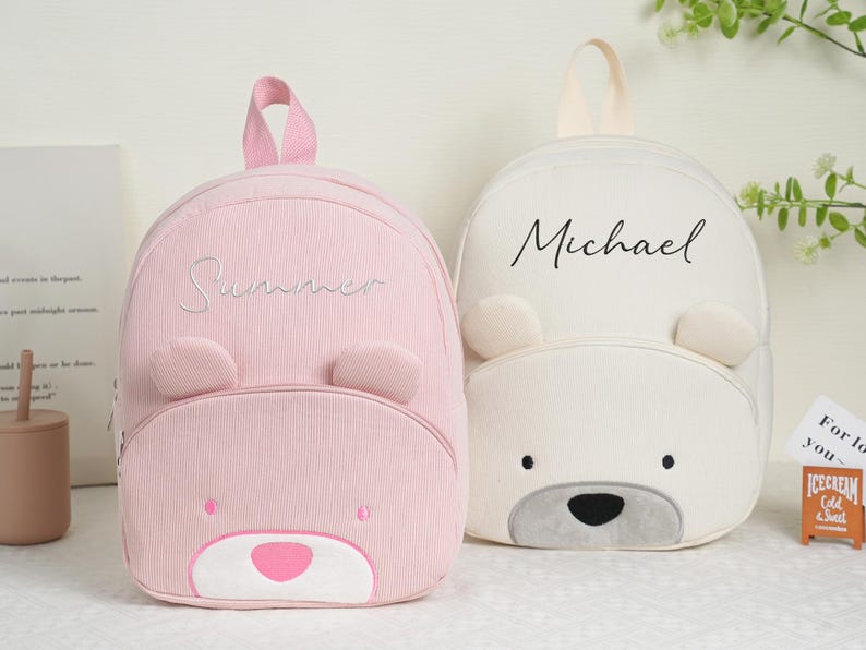 May include: Two children's backpacks, one pink and one white, each with a bear face design. The pink backpack has the name "Summer" embroidered on it, while the white one has "Michael". Both have bear ears and a top handle.