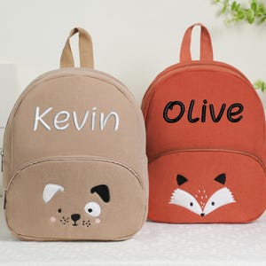 May include: Two corduroy backpacks. One is tan with "Kevin" embroidered in white and a dog face. The other is orange with "Olive" embroidered in black and a fox face. Both have top handles.