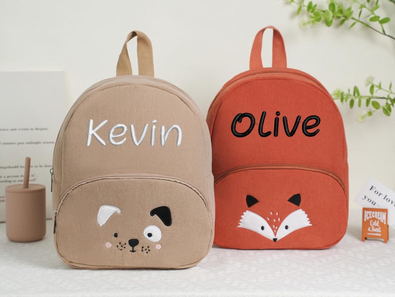 May include: Two corduroy backpacks. One is tan with "Kevin" embroidered in white and a dog face. The other is orange with "Olive" embroidered in black and a fox face. Both have top handles.
