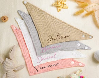 May include: Four cotton muslin baby bibs with different names embroidered on them. The names are Julian, Bonnie, Daniel, and Summer. The bibs are in shades of pink, beige, and gray.
