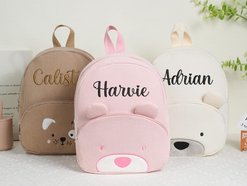 May include: Three personalized corduroy backpacks in tan, pink, and white. The tan backpack has a dog face design and the name "Calist" embroidered in gold. The pink backpack has a bear face and the name "Harvie". The white backpack has a bear face and the name "Adrian".
