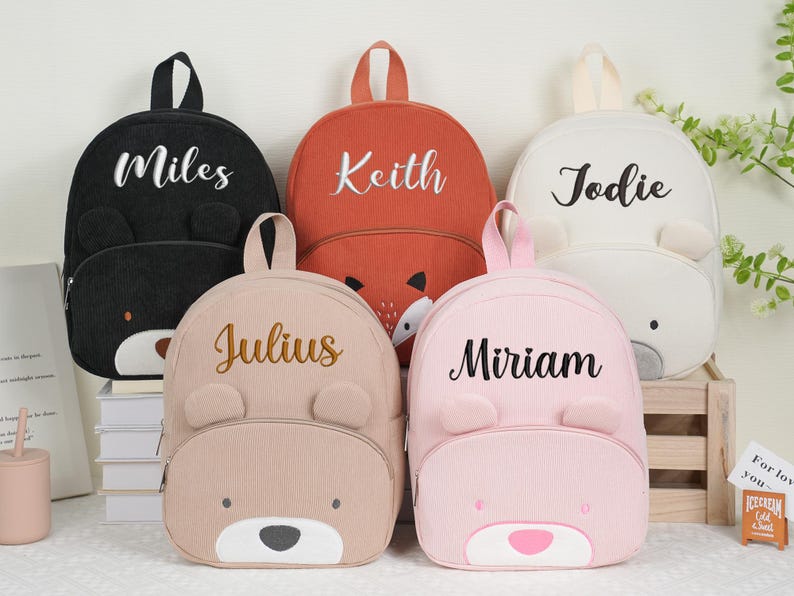 May include: Five corduroy backpacks in black, orange, white, tan, and pink, each with a bear face design and a name embroidered on the front. The names are Miles, Keith, Jodie, Julius, and Miriam.