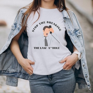 Puede incluir: Camiseta blanca con el texto "STOP THE BREAKING THE LAW A**HOLE" y un gráfico de una persona hablando por teléfono. La camiseta se lleva con una chaqueta vaquera y vaqueros azules.