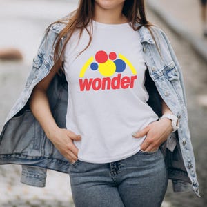 Pu&ograve; includere: T-shirt bianca con la scritta "wonder" in rosso e una grafica colorata sopra. La grafica include forme rosse, gialle e blu. La persona indossa una giacca di jeans e jeans blu.