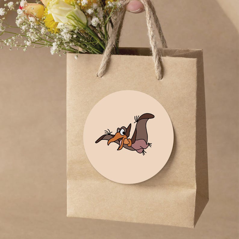 Pode incluir: Uma sacola de presente de papel marrom com al&ccedil;as de corda. Um adesivo redondo com uma ilustra&ccedil;&atilde;o de desenho animado de um pterod&aacute;ctilo &eacute; afixado na sacola. O pterod&aacute;ctilo &eacute; marrom, laranja e rosa. Um buqu&ecirc; de flores amarelas e brancas &eacute; vis&iacute;vel acima da sacola.
