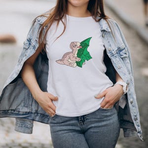 Pode incluir: Camiseta branca com um desenho de dinossauro de desenho animado e uma folha verde. O dinossauro &eacute; retratado em tons de castanho claro e rosa, com a l&iacute;ngua de fora. A folha &eacute; verde com um padr&atilde;o texturizado. Gola redonda.