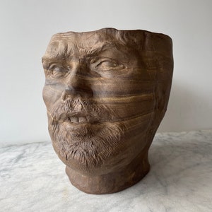 Op de afbeelding: Een bruine keramische plantenpot in de vorm van een menselijk gezicht met een baard. De pot heeft een getextureerd oppervlak en is ontworpen om planten in te houden.