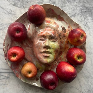 May include: A ceramic plate with a sculpted face in the center, surrounded by red apples. The plate has a white background with splashes of pink, orange, and brown.
