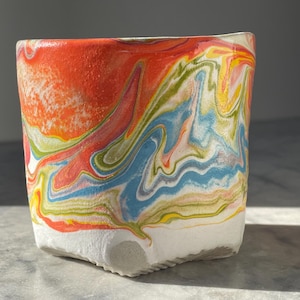 May include: A square ceramic planter with a vibrant, swirled design. The top is a textured orange, transitioning into a mix of blue, green, yellow, and white. The base is a solid white with a textured surface. A decorative piece for plants.
