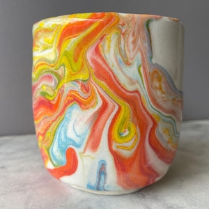 May include: A ceramic tumbler with a vibrant, swirled design in shades of orange, yellow, red, blue, and white. The cup has a cylindrical shape and a smooth, glossy finish, perfect for holding beverages.