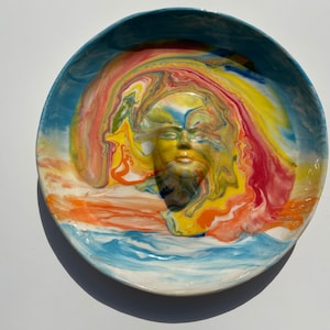 Puede incluir: Un plato de cerámica con un diseño abstracto colorido que presenta una cara en el centro. La cara está rodeada de remolinos de amarillo, naranja, rojo y azul.