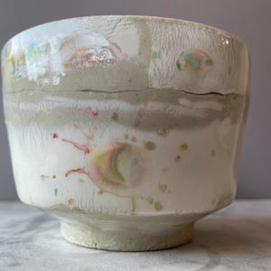 Może przedstawiać: Ręcznie robiona ceramiczna miska o szerokim, lekko nieregularnym kształcie. Misa ma białą podstawę, szary pas i teksturowaną, spękaną powierzchnię. Kolorowe, abstrakcyjne plamy i akcenty farby dodają wizualnego zainteresowania.
