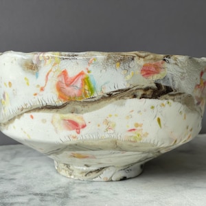 May include: A handcrafted ceramic bowl with a unique, textured surface. The bowl features a white base with colorful floral and abstract patterns in shades of red, yellow, and brown. The bowl is approximately 6 inches in diameter.
