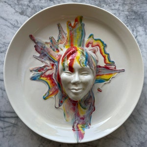 Puede incluir: Un plato de cerámica blanca con un rostro esculpido y detalles de pintura colorida. El rostro está centrado, con pintura de colores del arcoíris que irradia hacia afuera. El plato tiene un acabado liso y brillante.