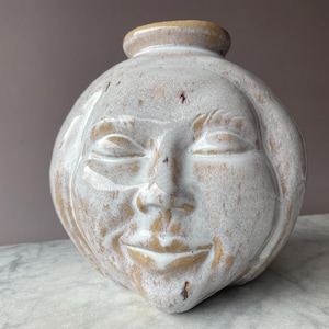 Op de afbeelding: Een keramische vaas in de vorm van een gezicht met gesloten ogen en een glimlachende mond. De vaas is geglazuurd in wit en beige met een gespikkelde afwerking.