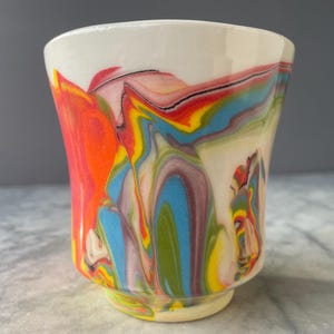 Puede incluir: Una taza de cerámica con un patrón jaspeado de remolinos vibrantes en rojo, amarillo, azul y verde sobre una base blanca. El recipiente tiene un acabado liso y brillante con un borde ancho que se estrecha hacia una base circular más pequeña.