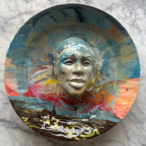 Puede incluir: Un plato de cerámica con un diseño abstracto colorido y una cara esculpida en 3D en el centro. La cara está pintada en tonos grises y marrones, y el fondo es una mezcla de azul, amarillo, rojo y naranja.