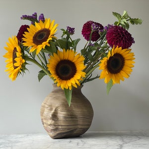 May include: A brown ceramic vase shaped like a face with a carved wood-like texture holds a bouquet of yellow sunflowers, burgundy dahlias, and purple flowers.