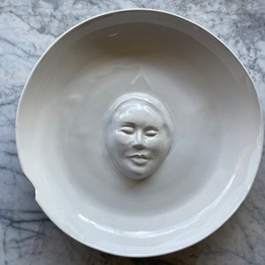 May include: A white ceramic plate with a raised, sculpted face in the center. The face has closed eyes and a neutral expression.