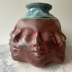 May include: A terracotta-colored ceramic vase featuring multiple sculpted human faces around its exterior. The vase has a wide, rounded base and a narrow neck finished with a glossy, textured teal-colored glaze at the top rim.