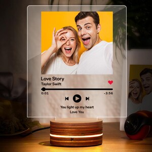 Può includere: Una luce notturna personalizzata con una foto di una coppia e il testo "Love Story" di Taylor Swift. Ha una base in legno e un pannello in acrilico trasparente con una grafica della canzone.
