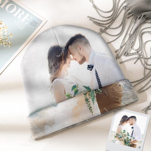 Könnte beinhalten: Hellgraue Mütze mit einem aufgedruckten Bild eines Paares, das sich umarmt. Das Bild umschließt die Mütze, mit einem braunen und grauen Band am unteren Rand. Ein kleines Foto des Paares ist ebenfalls vorhanden.