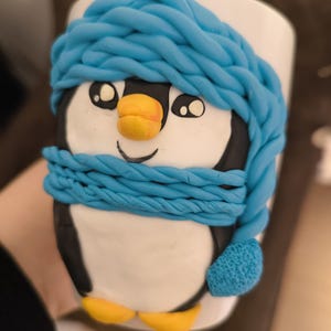 Könnte beinhalten: Eine weiße Keramiktasse, verziert mit einem 3D-Pinguin-Design. Der Pinguin hat einen schwarz-weißen Körper, gelbe Füße und einen Schnabel und trägt einen blauen Schal und eine blaue Mütze. Die Tasse ist ein skurriles und dekoratives Objekt.