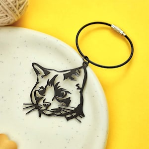 Könnte beinhalten: Ein schwarzer Schlüsselanhänger mit Katzenkopf. Der Schlüsselanhänger hat ein ausgeschnittenes Design eines Katzenkopfes mit Details wie Schnurrhaaren und Augen. Er ist an einer schwarzen, flexiblen Schlaufe mit Silberverschluss befestigt. Der Hintergrund ist gelb.