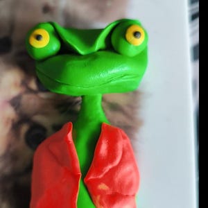 Könnte beinhalten: Eine skurrile, handgefertigte grüne Froschfigur mit großen Augen, jeweils mit gelbem Zentrum. Der Frosch trägt eine leuchtend rote Jacke. Dieser einzigartige, handgefertigte Artikel ist wahrscheinlich ein Dekorationsstück oder ein Sammlerstück.