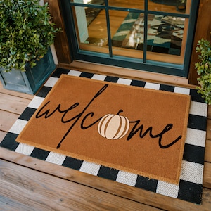 May include: A rectangular brown coir doormat featuring the word welcome in black cursive script, with a light orange pumpkin illustration replacing the letter o. The mat is layered over a black and white buffalo check patterned rug.