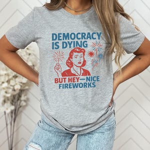 May include: A gray t-shirt with the text "DEMOCRACY IS DYING BUT HEY-NICE FIREWORKS" in blue and red. The graphic features a vintage-style illustration of a woman holding a sparkler, with fireworks in the background.