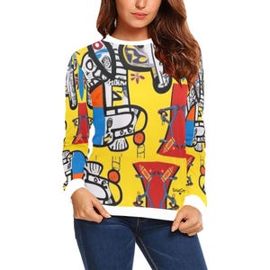 Peut inclure: Un t-shirt à manches longues avec un fond jaune et un motif abstrait coloré. Le motif présente des formes géométriques, des lignes et des figures en rouge, bleu et blanc.
