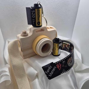 May include: A wooden camera with a yellow lens and a cream strap. Two black film canisters with yellow labels are attached. One canister is on top of the camera, and the other is next to a strip of film with photos. A black tag reads "Family Matters."