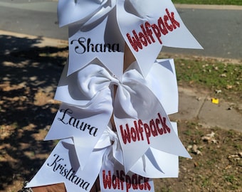 May include: White cheerleading bows with the names "Shana", "Laura", and "Kristiana" in black script. Each bow has a red glitter "Wolfpack" text. The bows are tied to a brown cardboard tube.