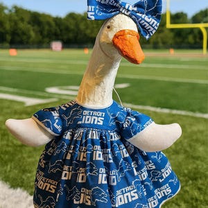 May include: A white goose statue wearing a blue dress and a matching bow, both featuring the Detroit Lions logo and text pattern. The goose has a textured white body and a bright orange beak, standing upright with its wings slightly extended outward.
