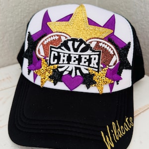 Puede incluir: Gorra de camionero en blanco y negro con un diseño "CHEER". La gorra presenta un panel frontal blanco con una estrella dorada con purpurina, estrellas moradas y balones de fútbol. La visera es negra y el texto "Wildcats" está en oro.