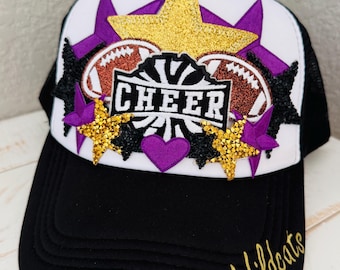 Könnte beinhalten: Schwarz-weiße Trucker-Mütze mit einem "CHEER"-Design. Die Mütze hat eine weiße Frontplatte mit einem goldenen Glitzerstern, lila Sternen und Fußbällen. Der Schirm ist schwarz, und der Text "Wildcats" ist in Gold gehalten.