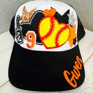 Könnte beinhalten: Schwarz-weiße Baseballkappe mit Mesh-Rückseite. Die Kappe hat eine weiße Vorderseite, die mit einem gelben Softball-Herz, orangefarbenen Sternen und der Zahl 9 verziert ist. Der Name "Gwen" steht in orangefarbener Schrift auf dem Schirm.