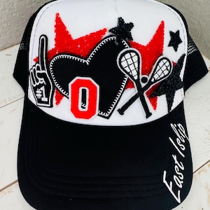 Puede incluir: Gorra de camionero en blanco y negro con un panel superior blanco con explosiones de estrellas rojas, un corazón negro y palos de lacrosse. La gorra tiene el número 1, la letra O y las palabras "East Islip" en blanco.