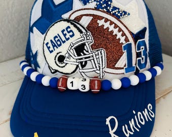 Può includere: Un cappellino trucker blu e bianco con una toppa a forma di casco da football americano con la scritta EAGLES, un pallone da football marrone glitterato, stelle blu e bianche e il numero 13. Un bordo di perline con il numero 13 e la scritta Runions.
