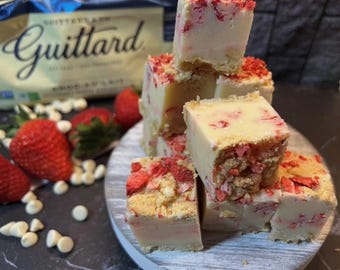 Può includere: Quadrati di fudge al cioccolato bianco ricoperti con pezzi di fragole secche. Il fudge è disposto su un tagliere di legno bianco con gocce di cioccolato bianco sparse intorno.